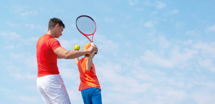 A young tennis coach or instructor teaches a child tennis player service techniques. Kids tennis on the court. Tennis school or club. Blue sky background. Banner size. Copy space for text.