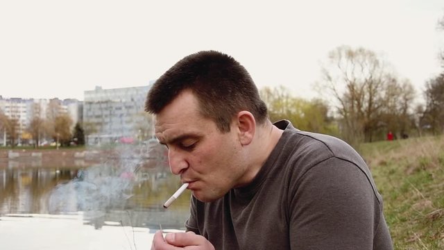 A man lights a joint in nature against the background of houses and a lake, takes a deep drag on the smoke and exhales, medium plan.