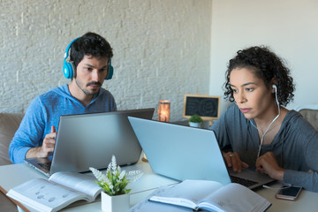 Focused Young freelancer couple with headphones working and typing in laptop. Inside the house in iving room. Freelance, home office, remote work, online concept.