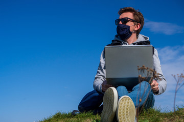 Naklejka na ściany i meble A young man in dark glasses and medical mask sits on grass in Park with laptop in his hands against blue cloudless sky. Remotely works during allergies or coronavirus pandemic. office outdoors