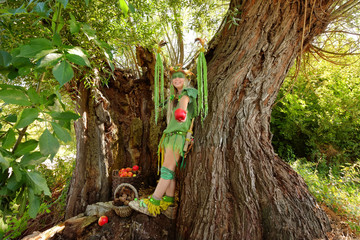 A young girl is dressed up as a fairy.
She is seen inside a hollow tree with
a basket full of red apples.