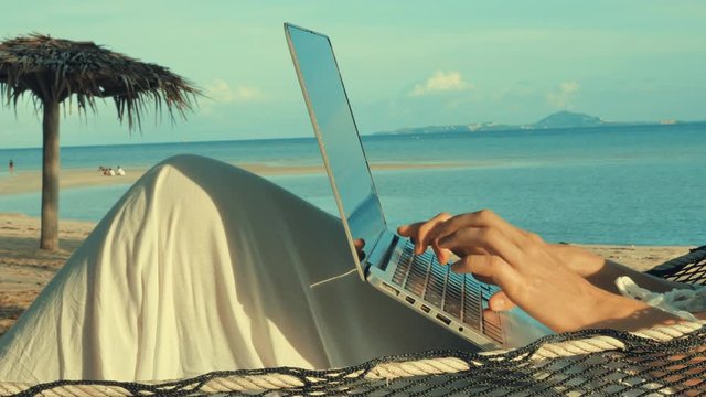 Young woman working on laptop lying in hammock at sand beach of tropical island. Freelance outdoor work concept