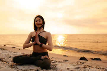 Fototapeta na wymiar A young girl sits in a lotus position on the seashore at sunset. Girl practices yoga by the sea