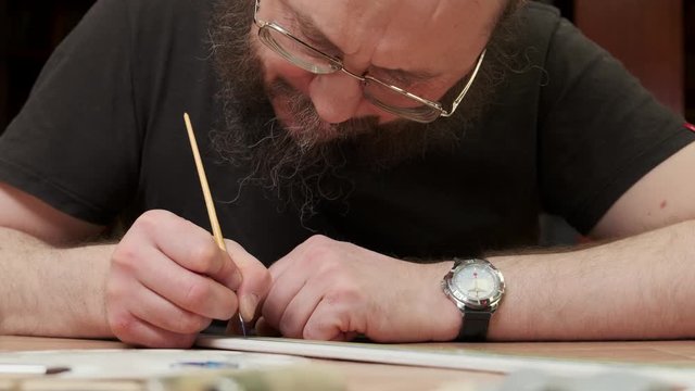 bearded person with glasses paints passenger jetliner model at table in Sunday school class closeup slow motion