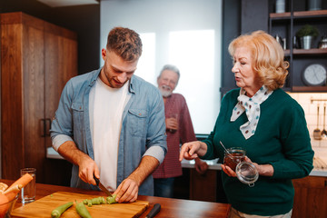 Young man is chopping peppers with kitchen knife, while his mother standing next to him with jar full of coffee. Father is standing next to the kitchen window.