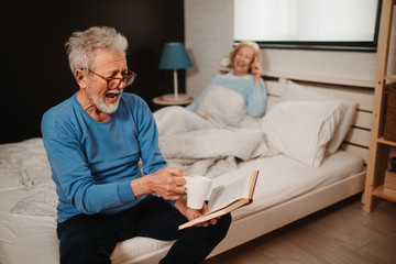 Close photo of laughting elderly man with grey hair and beard and with glasses reading book while drinking morning coffee. Blonde woman is talking over the phone while lying in bed behind him.