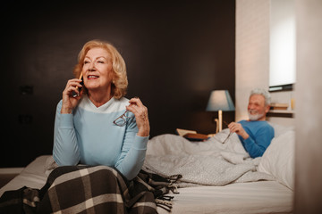 Portrait of blonde elderly woman having phone call with dear person. Her husband smiles while lying behind her in bed.