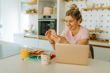 Portrait of young woman with wireless headphones in her ears looking at her smartphone. Blonde woman is sitting in front on laptop while sitting in the kitchen.