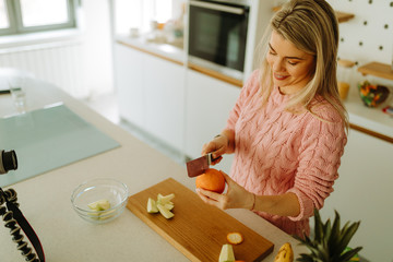 Portrait of blonde woman in pink sweater peeling an orange for fruit salad that she is making for breakfast. Food blogger is recording new video clip for her blog.