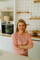 Photo of young blonde woman with beautiful smile in pink sweater posing with crossed arms in front of camera in kitchen at home.