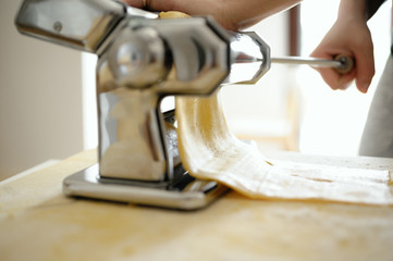 Staying at home with your family and preparing fresh home-made pasta (tagliatelle): mom passing the dough in the rollers with pasta machine on a wooden board.
