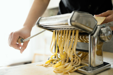 Staying at home with your family and preparing fresh home-made pasta (tagliatelle): mom cutting sheets of pasta with pasta machine on a wooden board.