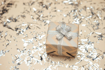 Gift box wrapped in kraft paper with silver ribbon and bow on beige sparkling background. Holidays present concept. Christmas background, festive decor. 