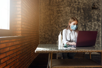 young blonde girl in a white blouse and a medical mask sits in a room at a table with a laptop and medicines