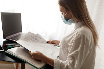 young blonde girl in a white blouse and a medical mask works in the room with documents and a laptop. Work at home.