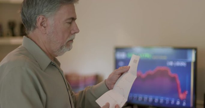 A television with stock market graphs and charts in the background a mature man looks at his investment statement which contains nothing but bad news because of COVID19 pandemic.
