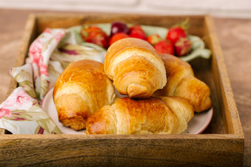 Freshly croissant in wooden tray, fresh berries, napkin, brown background. Close up