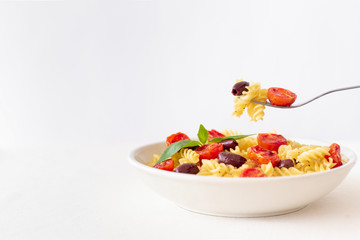 Pasta fusilli with baked cherry tomatoes, kalamata olives and basil on white plate with fork holding pasta. Copy space on the left.