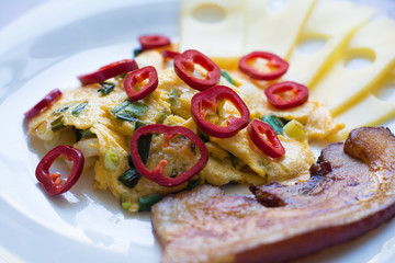 American style scrambled eggs with diced green onions, garnished with sweet red pepper slices, and served with one thick pan-fried smoked bacon strip, and pieces of Emmental cheese.