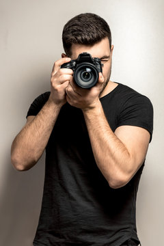 young male photographer in a black t-shirt stands with a camera on a gray background. Vertical frame