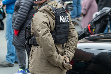 A male police officer stands on a street among a crowd of people with his hands behind his back. He is wearing a bulletproof vest with white lettering spelling police. There's a black car nearby