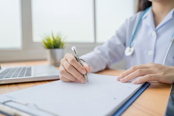healthcare and medical care working from home concept, beautiful female Asian doctor working in home office smiling writing one clipboard diagnosing patients health using computer laptop and tablet