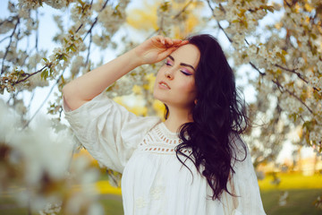 Incredibly tender girl in blossoms, on a background of a tree with white flowers, a model in a white boho dress. Brunette with long hair in the spring. Girl with a pierced nose. 