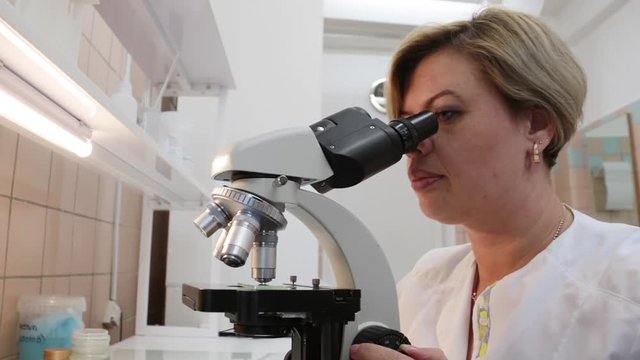 Beautiful girl nurse carefully examines bacteria through a microscope. Close-up. The camera moves smoothly. Soft white light.