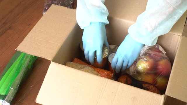 A Volunteer In Warehouse Collects Donation Food Box For A Family In Need During The Covid-19 Period. Aid Help And Charity Help In The Financial Crisis. Protective Face Mask And Hands With Gloves