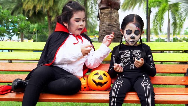 Asian kids play together with Halloween costumes and makeup ghost style, children have fun knock door for tric or treat share sweet together, primary school student sit in park after Halloween
