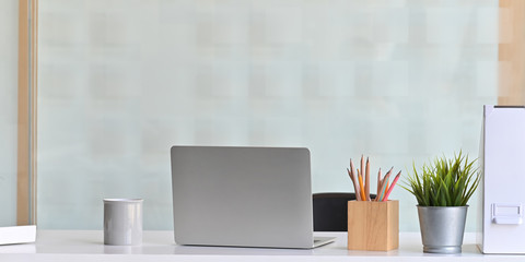 Photo of Computer laptop that putting on white working desk and surrounded by coffee cup, document file, wooden pencil holder and potted plant. Orderly workspace concept.