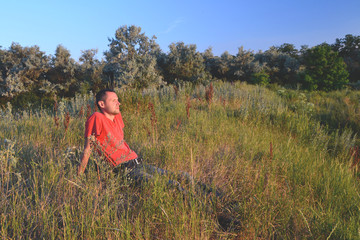 Naklejka na ściany i meble tired lonely young man in a red t-shirt and jeans relaxig in grass in the meadow near the trees at sunset 