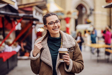 Smiling young woman standing on the street on chilly weather and holding disposable cup with coffee.