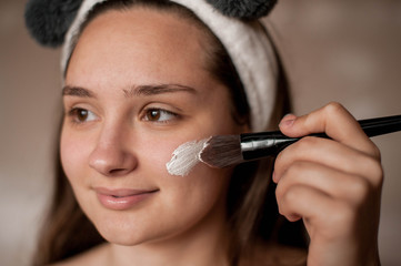 a brunette girl without makeup with problematic skin with a bandage with ears on her head naked wrapped in a towel after a bath doing Spa treatments applies a white clay mask to the face with brush
