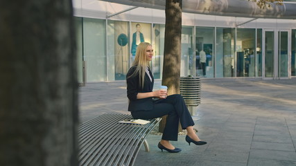 Elegant modern woman sitting on bench and waving with hand to friend while speaking on smartphone outdoors