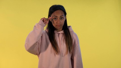 Attractive black woman twisting her index finger at temple on yellow background. She looking at camera with disagreement.