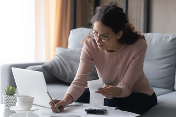 Focused millennial woman in eyewear writes out information from invoices, calculating monthly...