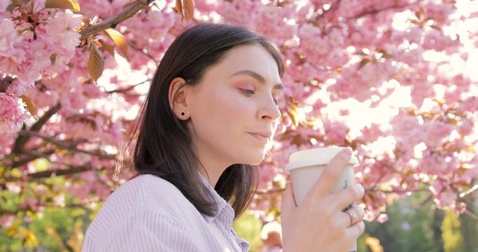Charming lady drinking coffee and resting in blooming garden at sunny day. Young girl enjoying takeaway coffee from eco mug while standing under flowering cherry tree in beautiful city park