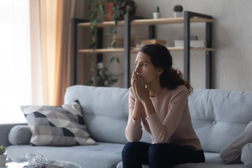Thoughtful doubtful young woman sitting alone on couch, feeling nervous about hard decision....