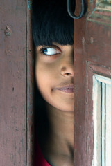 Indian rural child is peeping in the door. Portrait of a little girl is looking from the door. Beautiful eye of a child on black background. Dramatic look of a little girl in India.