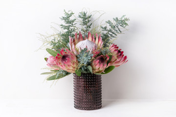 A elegant flower arrangement of a pink king protea, pink ice proteas and wattle leaves, in a purple glass vase on a table, with a white background.  - obrazy, fototapety, plakaty