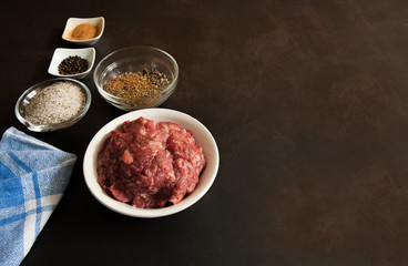 Raw fresh homemade ground beef in a white bowl, salt pepper and spices on a dark concrete background. Selective focus. Free space.