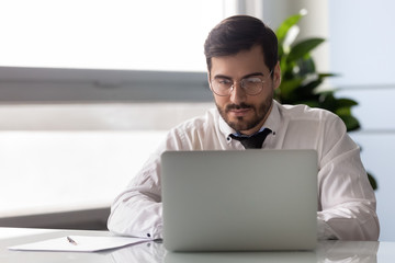 Young serious businessman working at laptop in office. Male employee reading business mail at computer. Worker man conduct electronic transaction, guy using modern technology.