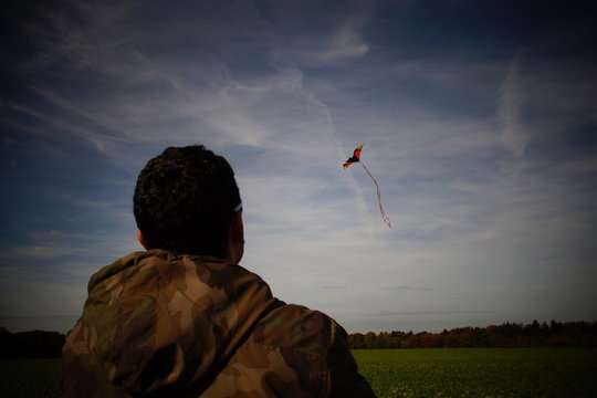 Rear View Of Army Soldier Looking At Kite On Grassy Field Against Sky
