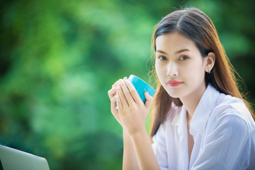An Asian woman holds a coffee cup for drinking and she uses a notebook computer to work at home in the morning.