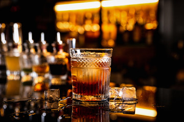 A cocktail in an old fashioned glass on a bar counter with a reflection, ice cubes around, bottles...