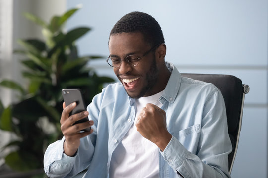 Happy african american businessman with glasses celebrating business success achievement, shouting. Diverse employee feeling motivated by good work looking at smartphone screen, receive great news in