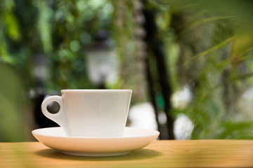 White coffee mug on wooden table in the tropical garden background with a refreshing morning light.