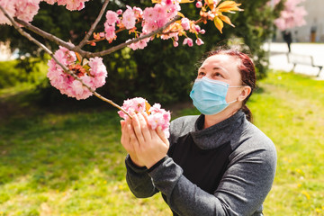 The girl in the park sniffs a sakura flower in a protective mask. A woman walks in a sakura park.Outdoor.