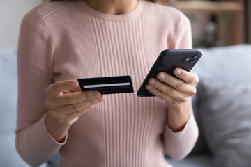 Close up young woman holding plastic credit card in one hand and telephone in another, entering cvv...
